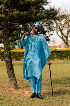 Person in vibrant blue traditional African clothing posing with a cane outdoors.