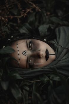 A woman with dramatic tribal makeup lies in dark foliage, creating a mystical vibe.