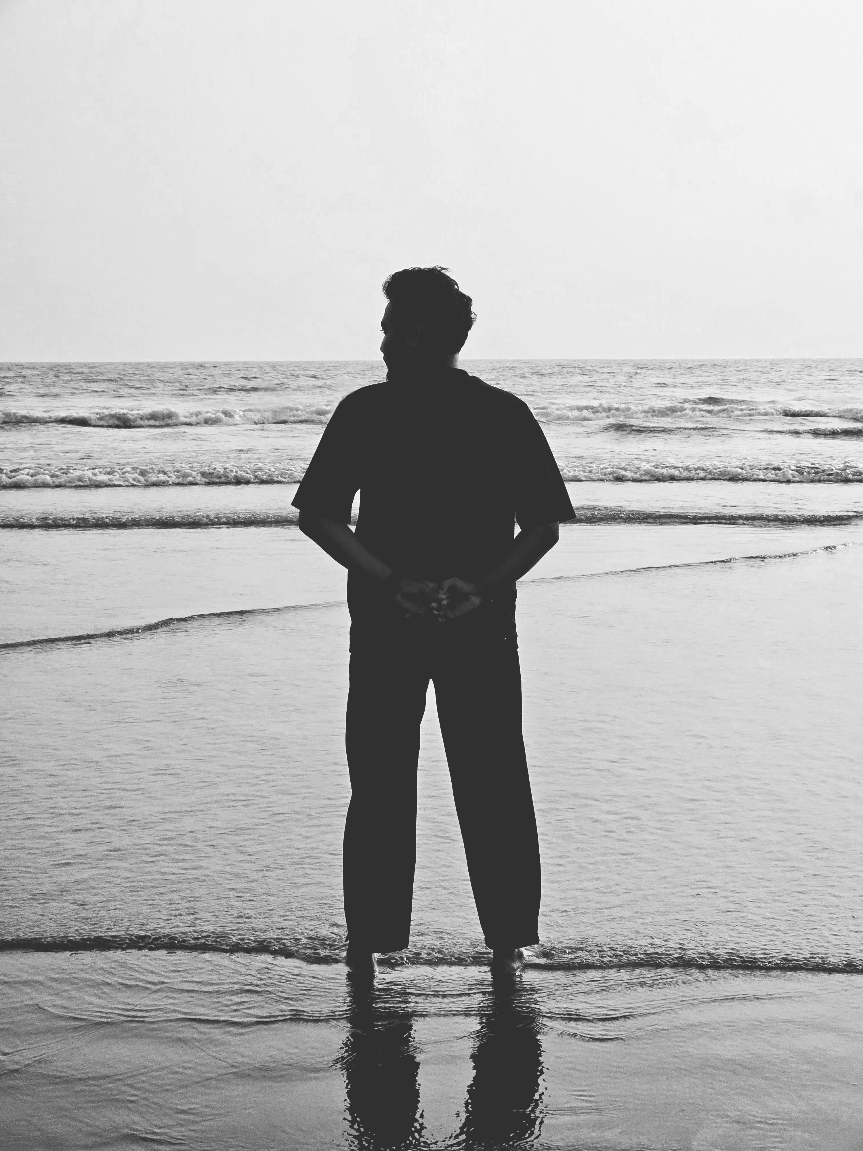 A silhouette of a man standing at the edge of the ocean shore. Reflective moment captured in black and white.