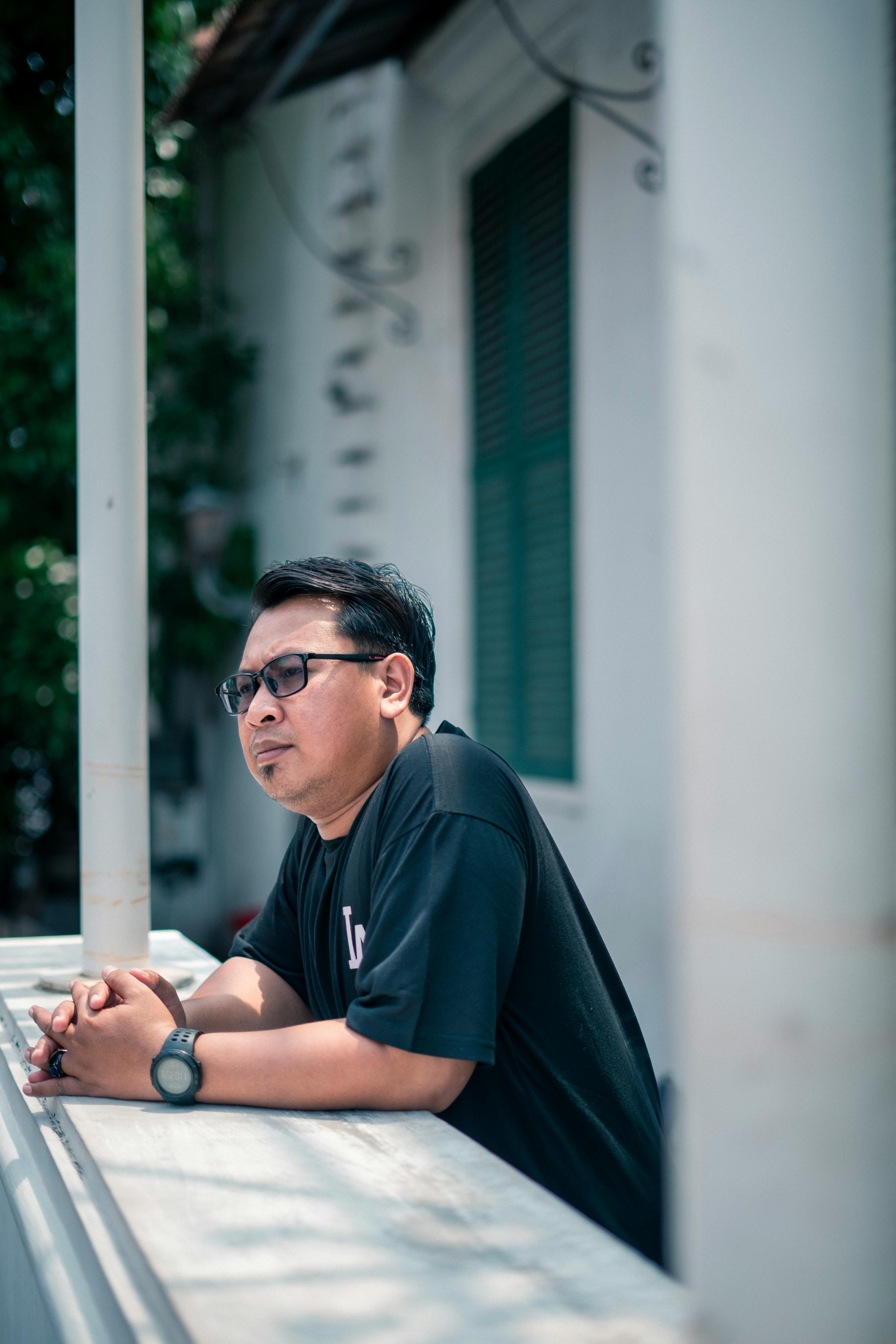 An Indonesian man in glasses daydreaming on a Jakarta balcony, captured in natural daylight.