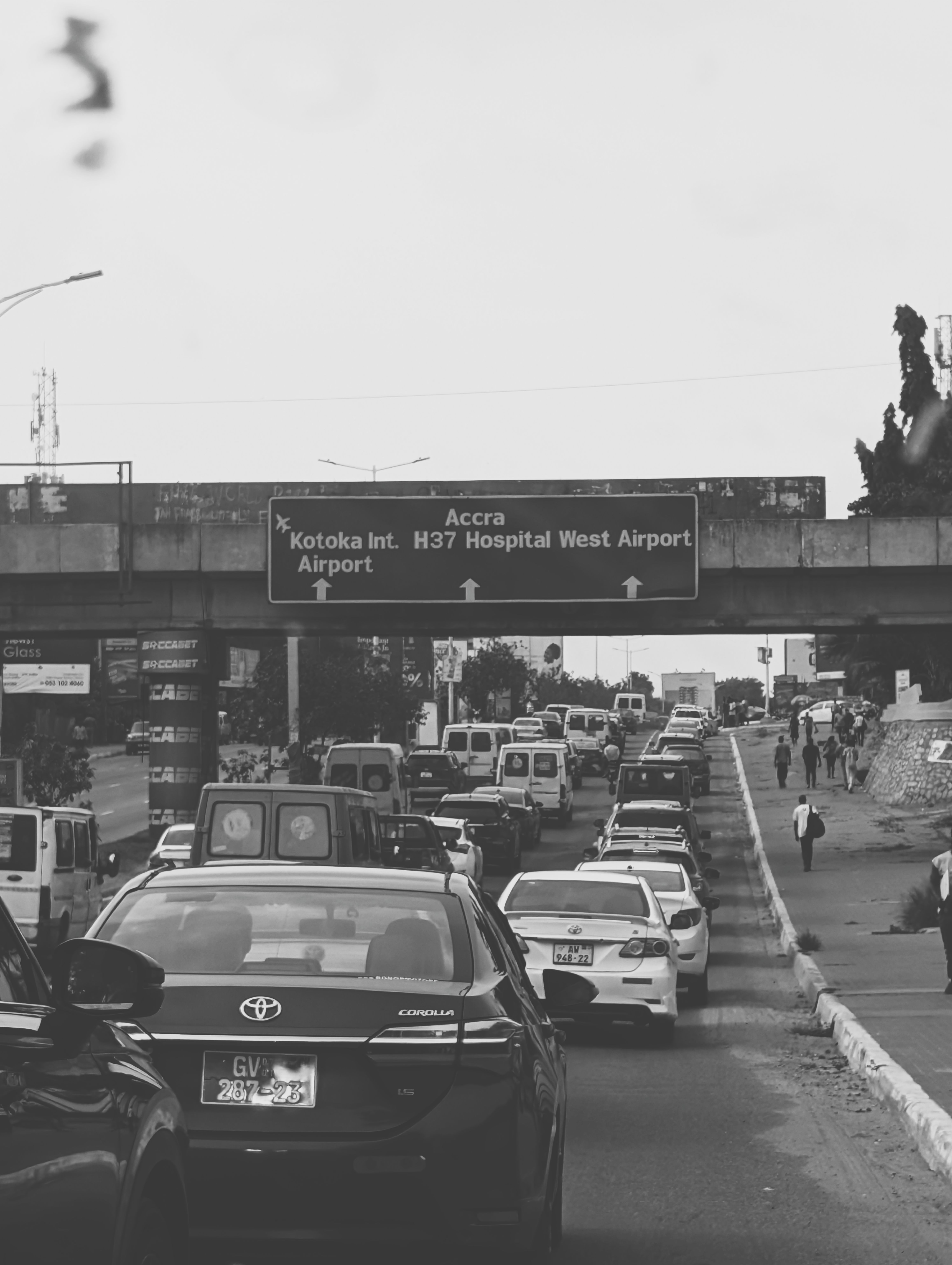 Black and white photo of heavy traffic on a road in Accra near a sign for Kotoka International Airport.