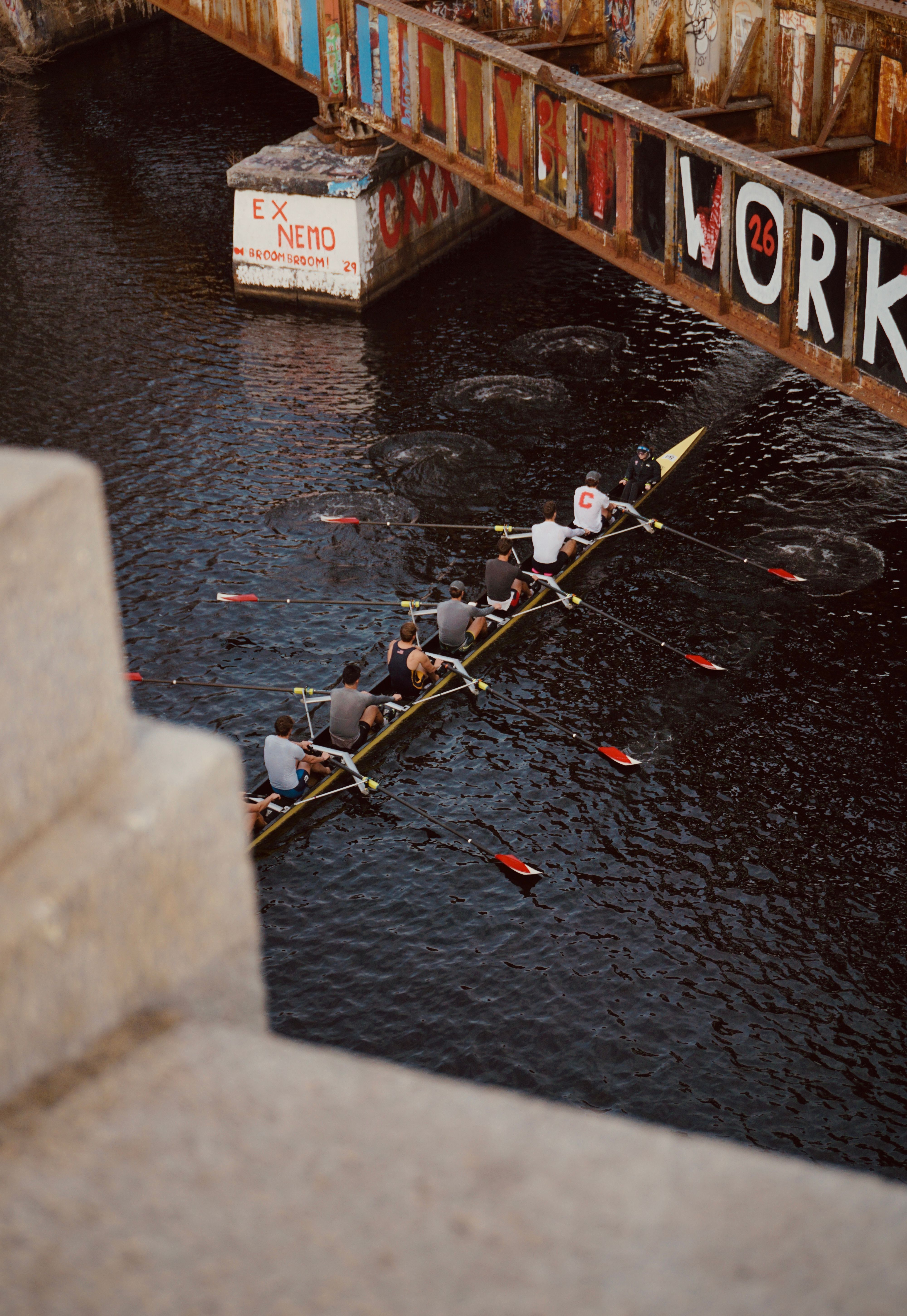 A rowing team navigates a waterway beneath a graffiti-covered bridge.