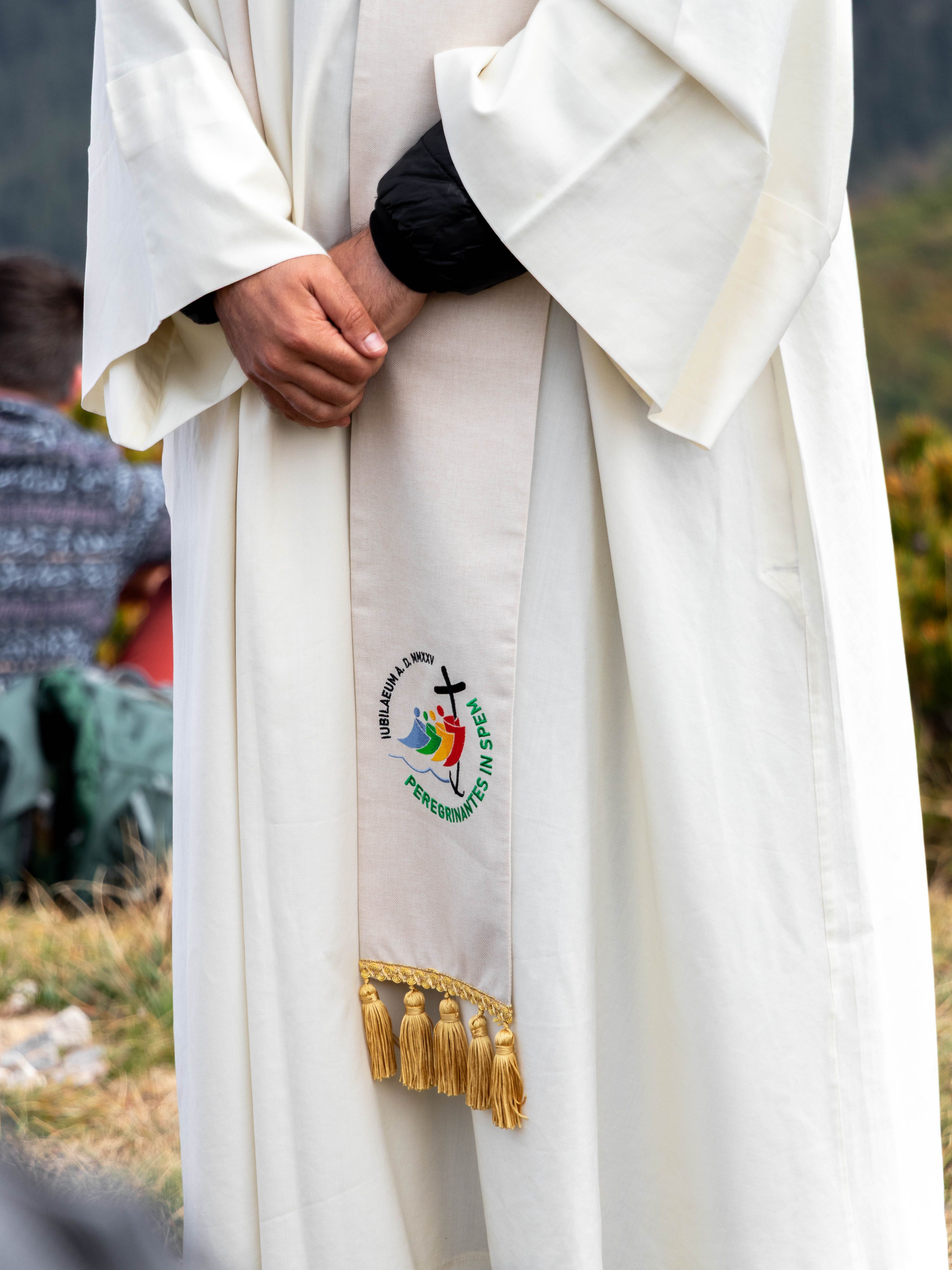 A cleric in a white robe stands outdoors, featuring an embroidered religious symbol.