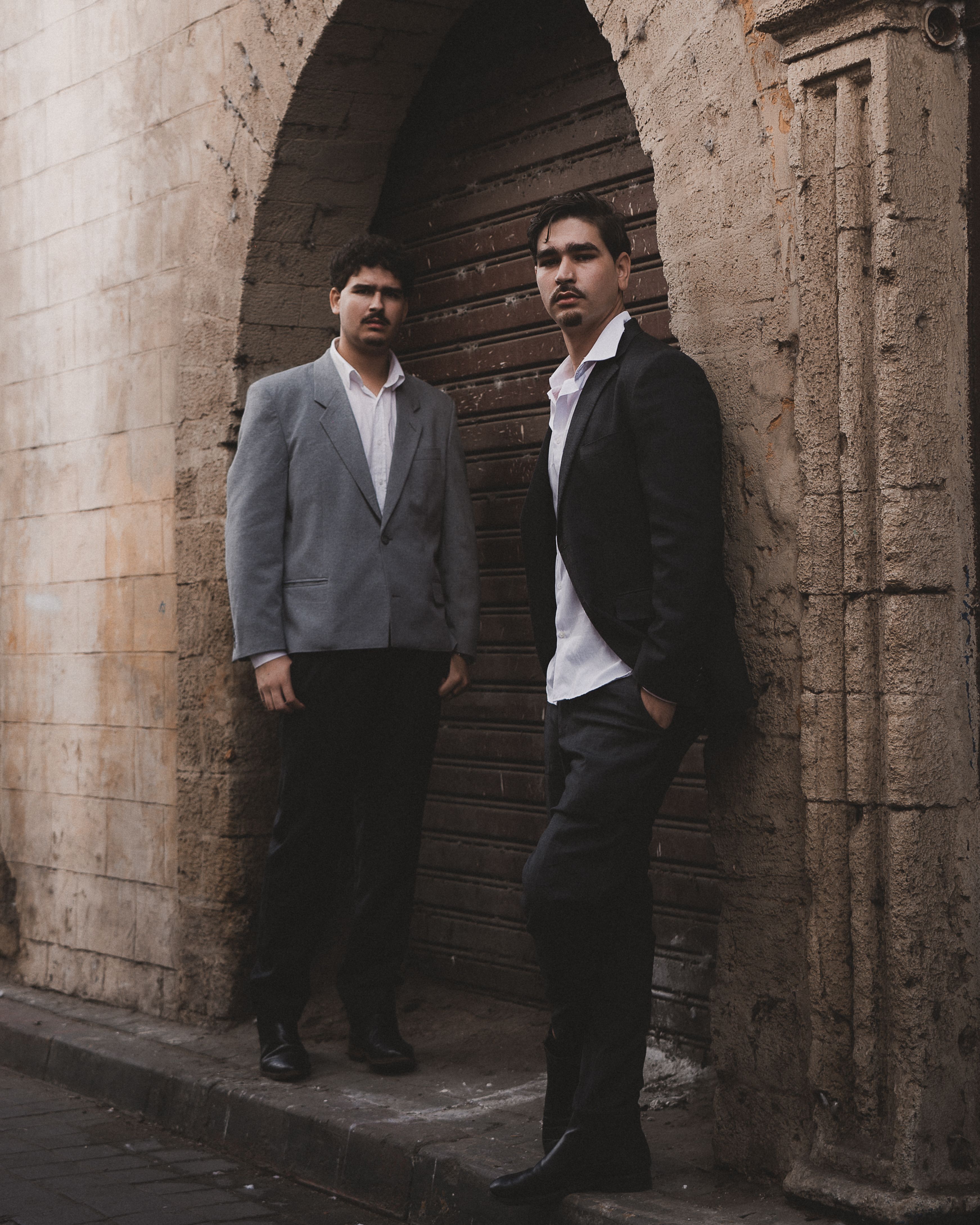 Two men in formal wear pose stylishly by a rustic archway in Casablanca, Morocco.