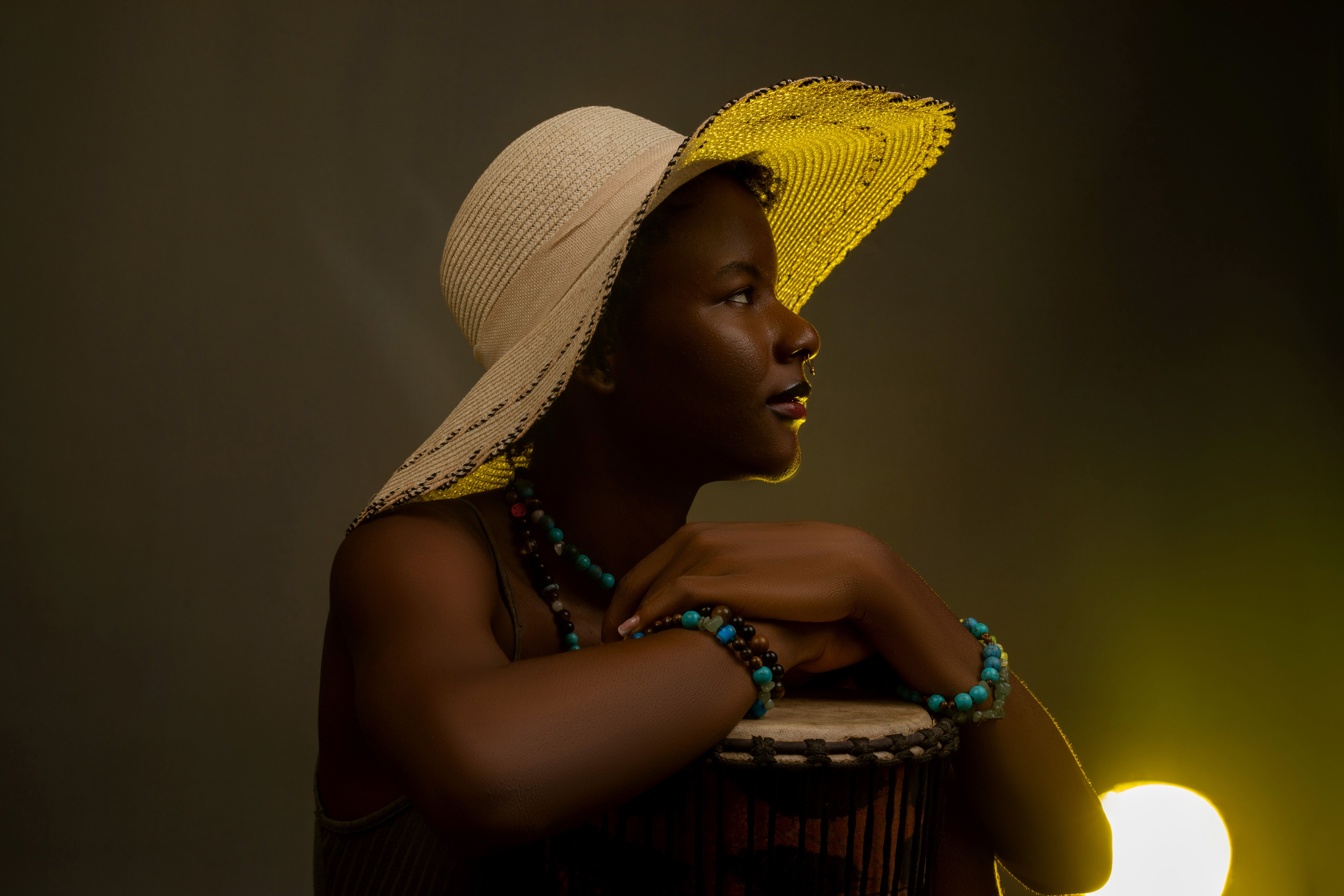 Portrait élégant d'une femme coiffée d'un chapeau de paille et portant des perles, appuyée sur un tambour, dans une lumière chaude.