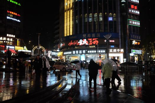 A bustling city street at night with neon lights and people under umbrellas on a rainy evening.