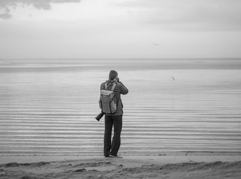 A lone photographer taking a scenic shot at a tranquil beach with overcast skies, in monochrome tones.