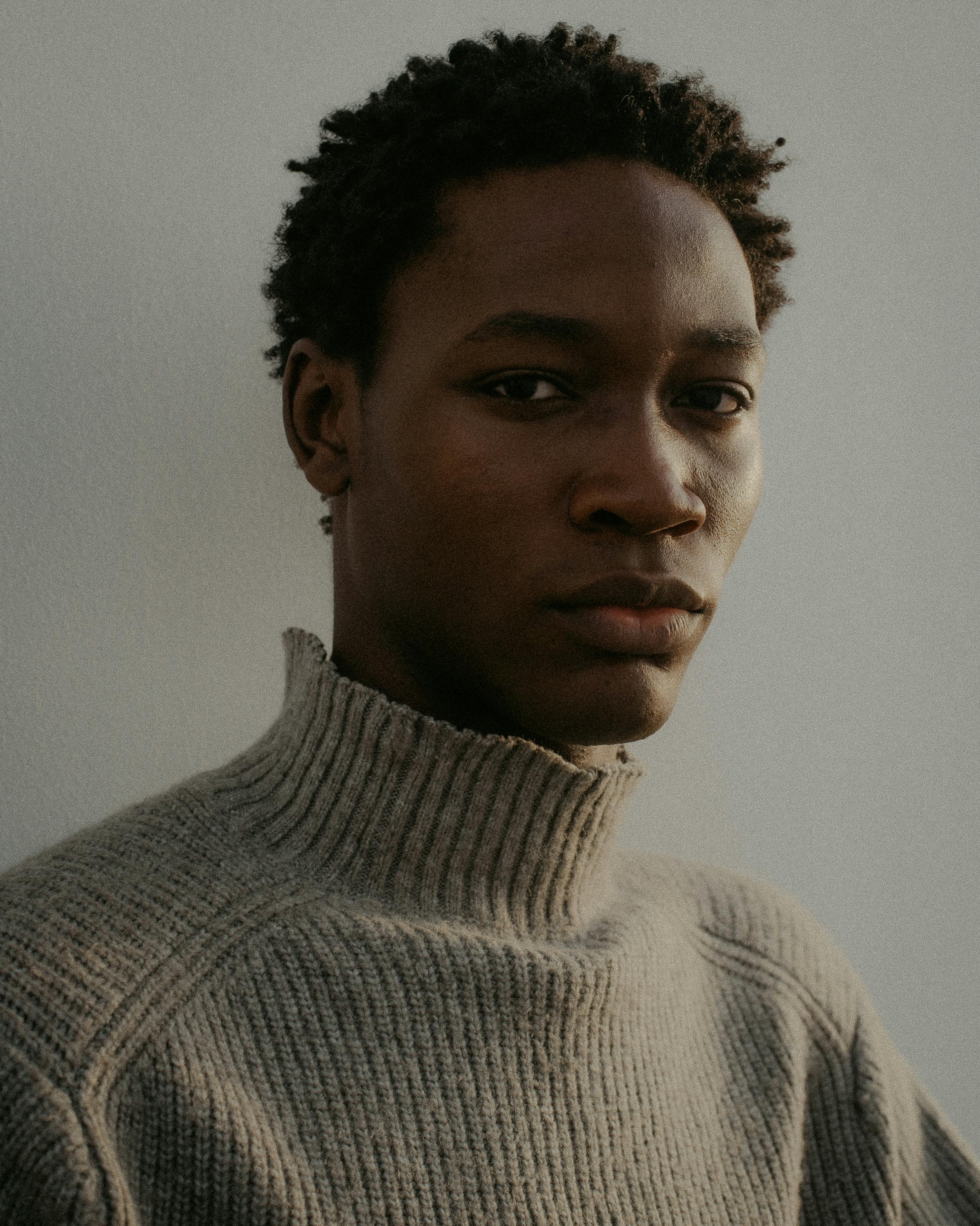 Moody portrait of a young black man wearing a turtleneck sweater with a neutral expression.