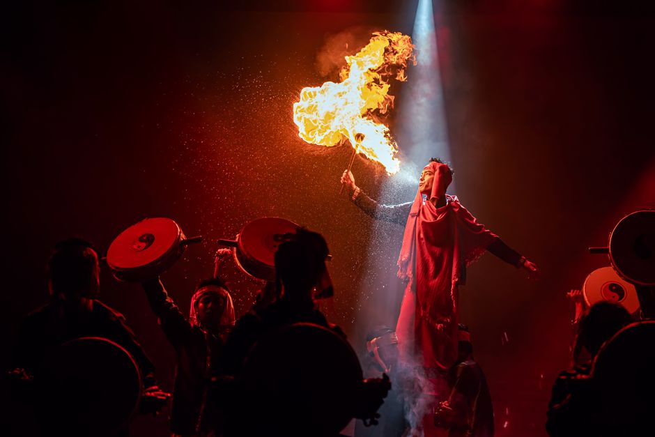 A mesmerizing fire performer on stage surrounded by drummers in a dynamic, high-energy show.