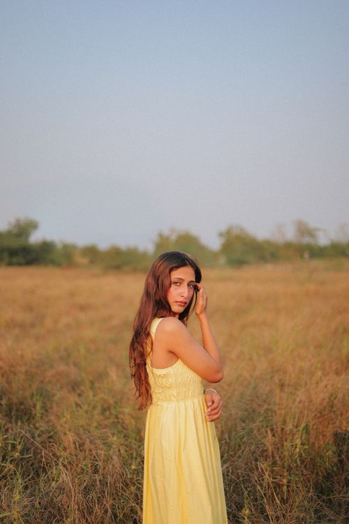 Free A young woman in a yellow dress stands in a vast open meadow under a clear sky. Stock Photo