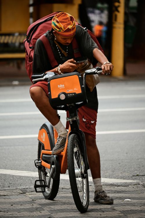 Free Man on orange bike rental stops on street while using smartphone, balancing urban life and connectivity. Stock Photo