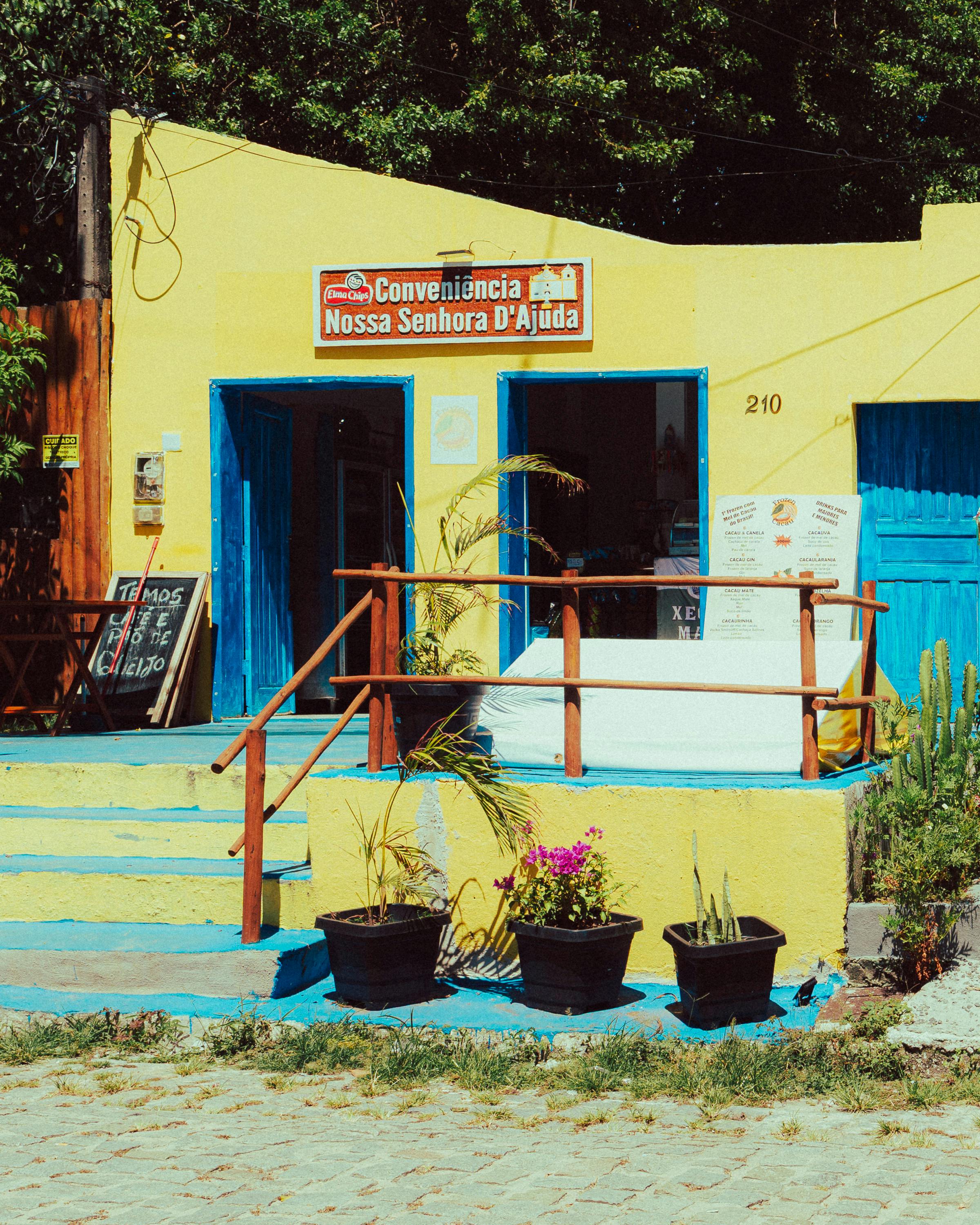 A vibrant yellow convenience store with blue accents set in lush green surroundings, captured in sunlight.