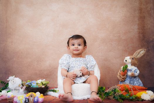 Cute toddler sitting between Easter decorations, holding a stuffed toy, surrounded by colorful eggs and bunny figures.