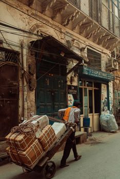 A vendor pulls a cart loaded with goods through a busy street with old buildings and signage.
