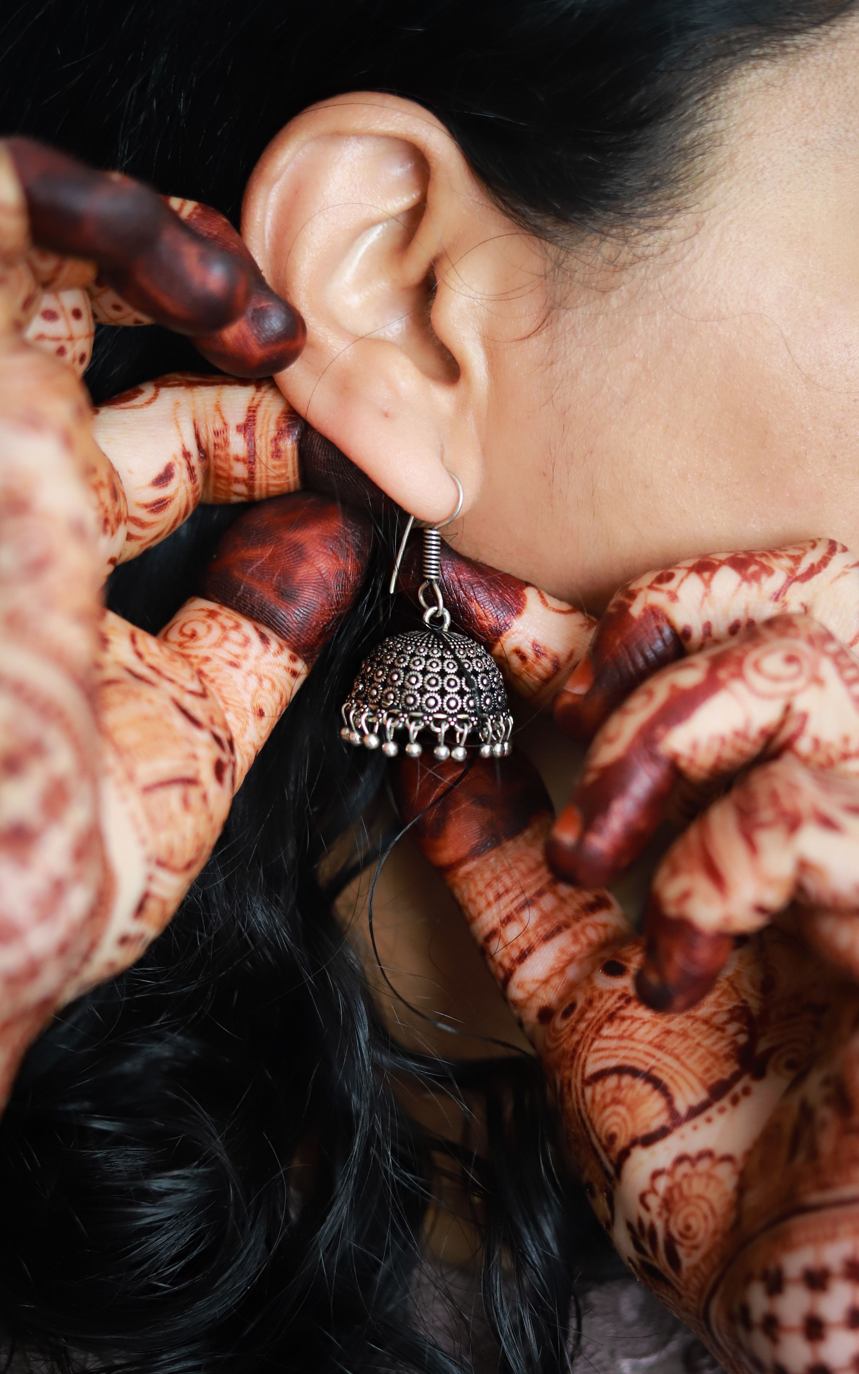 Detailed shot of henna-decorated hands adjusting a traditional jhumka earring on a woman's ear.