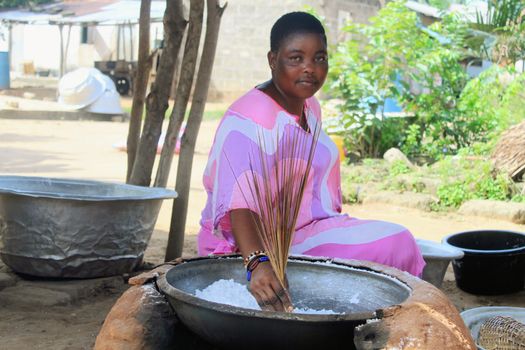 A woman in traditional attire preparing food in an outdoor setting with a large pot.