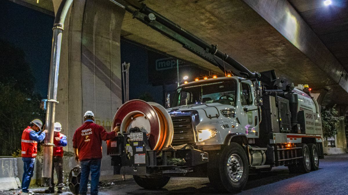 Workers operate a vacuum truck under a bridge at night for drain maintenance