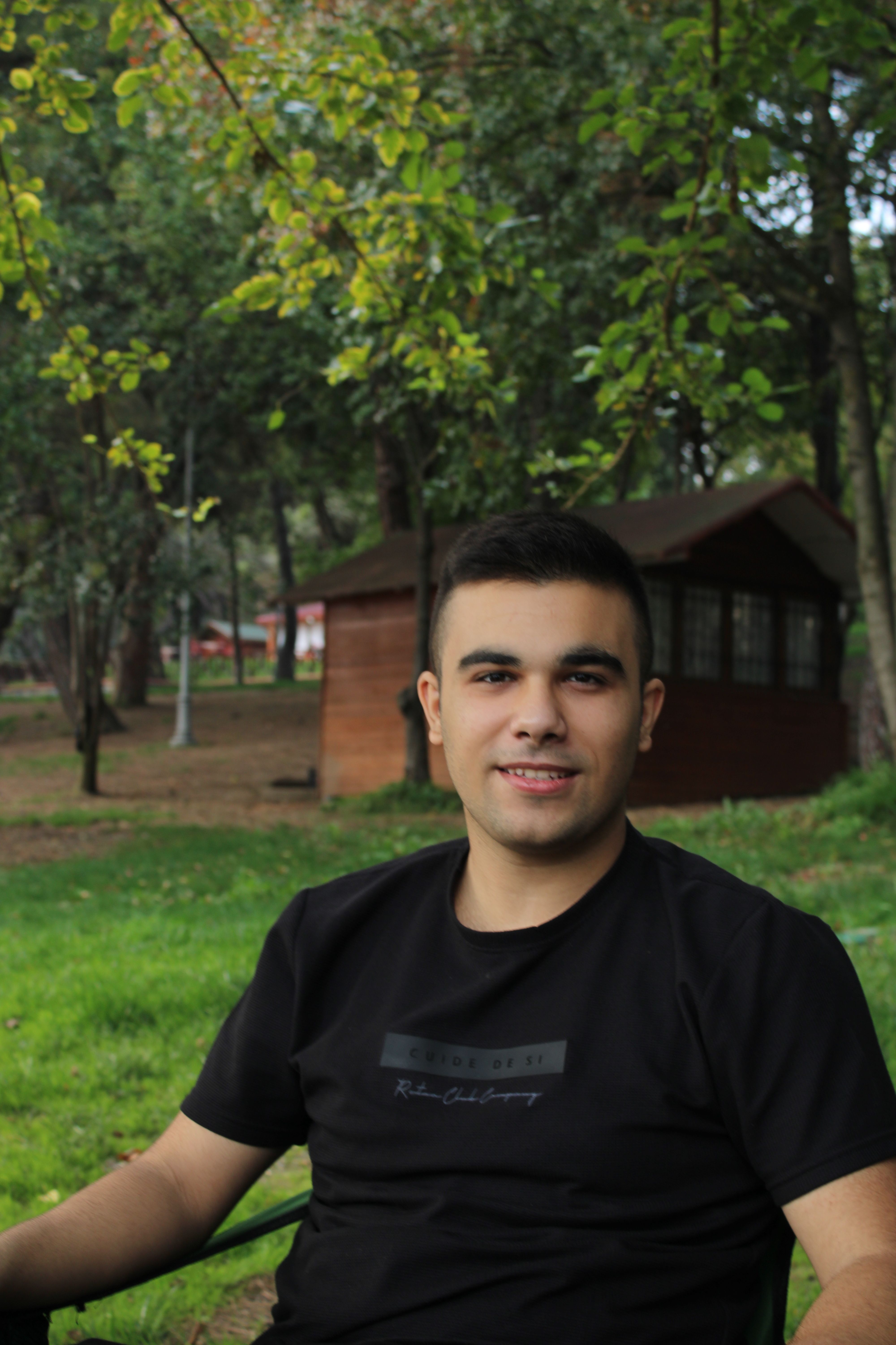 Portrait of a young man smiling while seated outdoors among trees in a park.