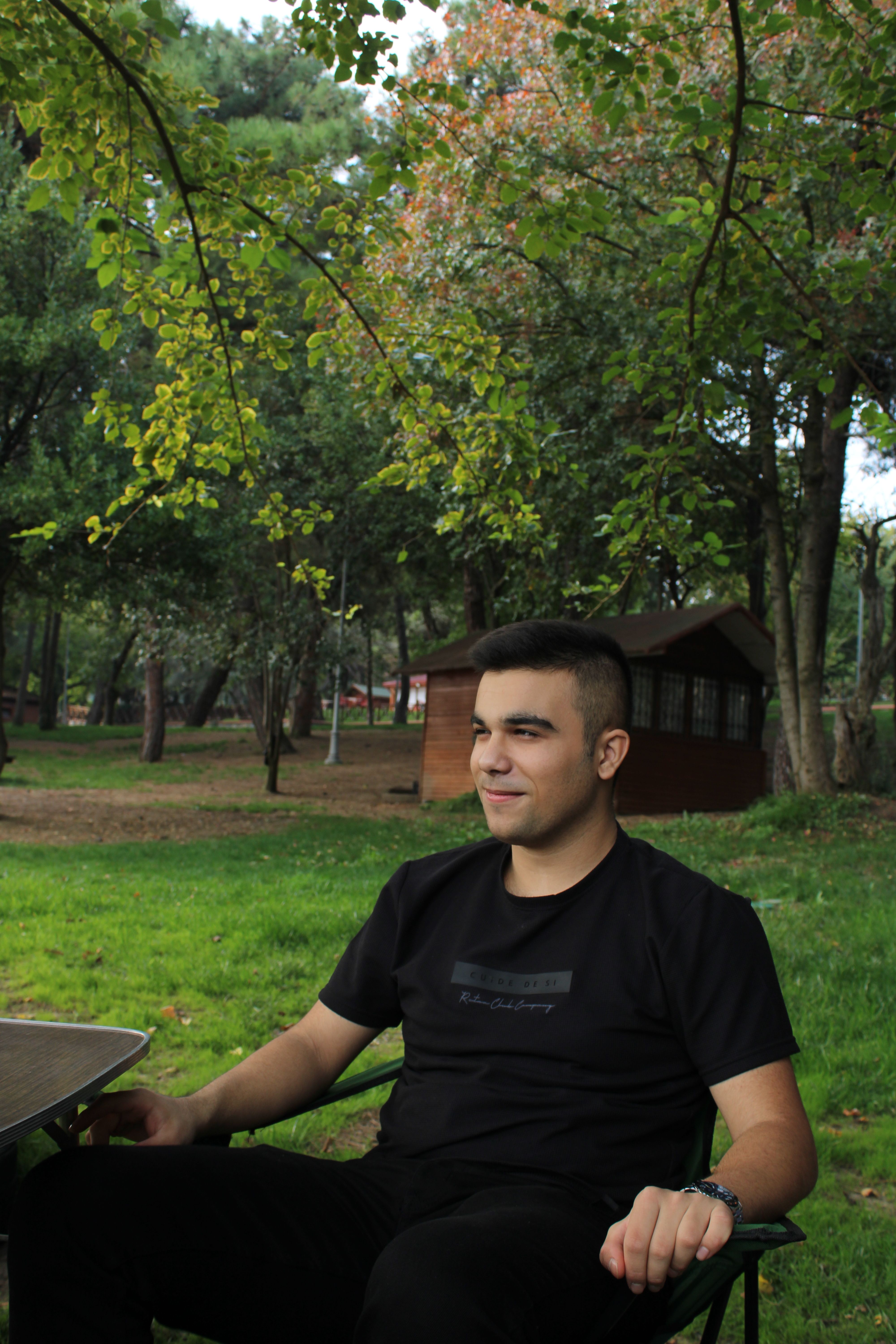 A young man enjoying a peaceful afternoon outdoors, seated by a wooden cabin in a lush green park.