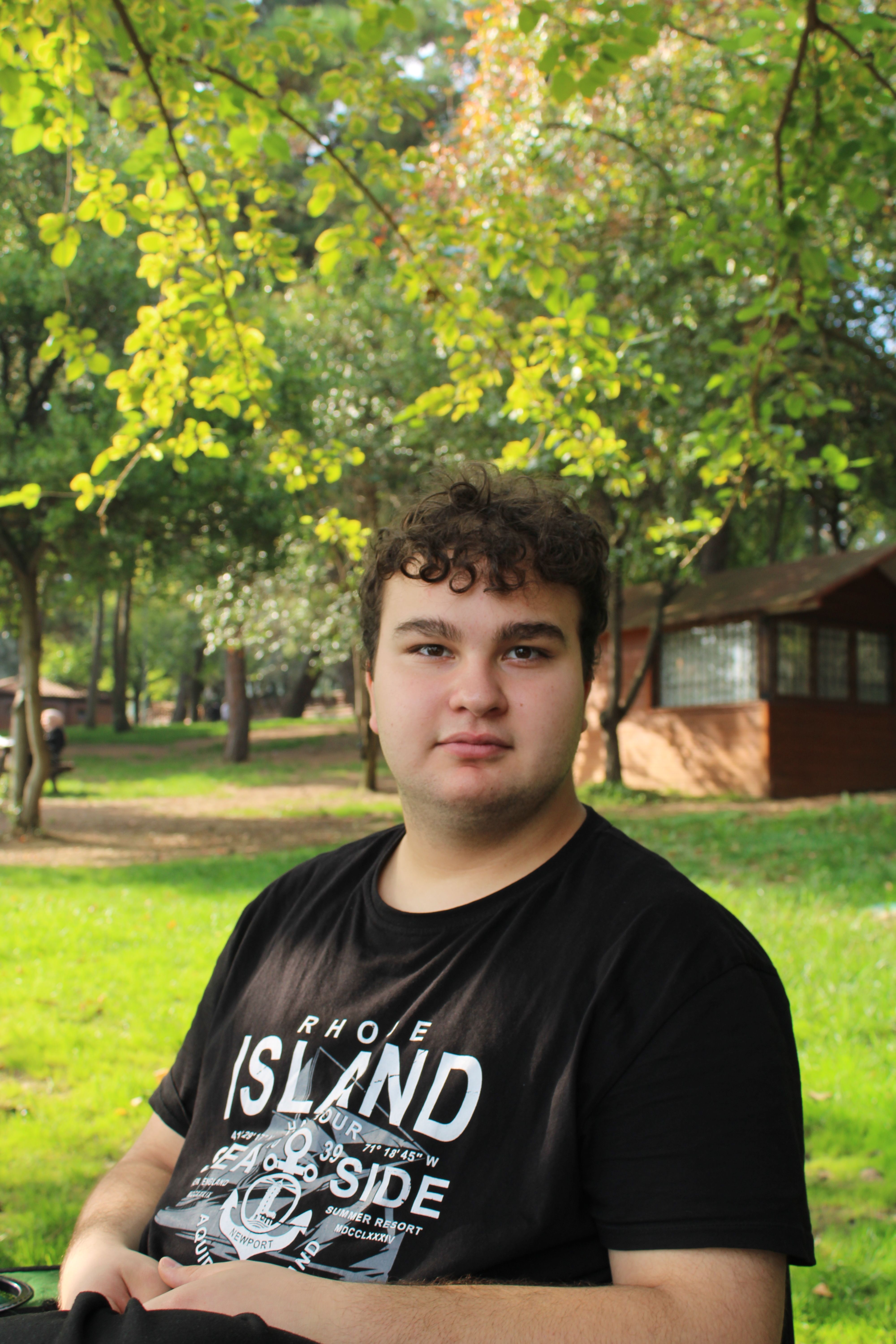 A young man sits calmly in a sunny park, surrounded by lush greenery and wooden buildings.