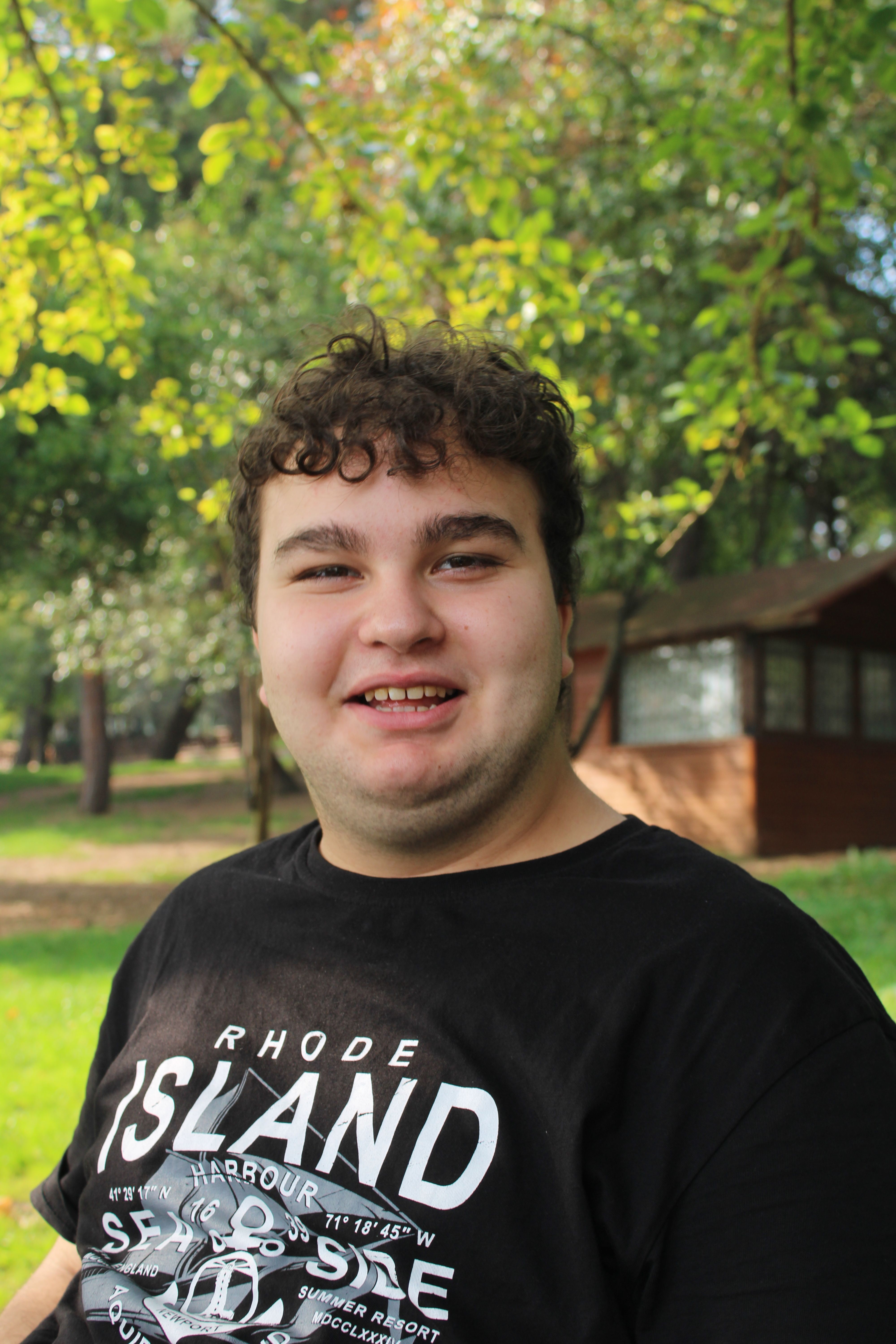 Portrait of a smiling young man wearing a Rhode Island T-shirt, outdoors in a sunlit park.
