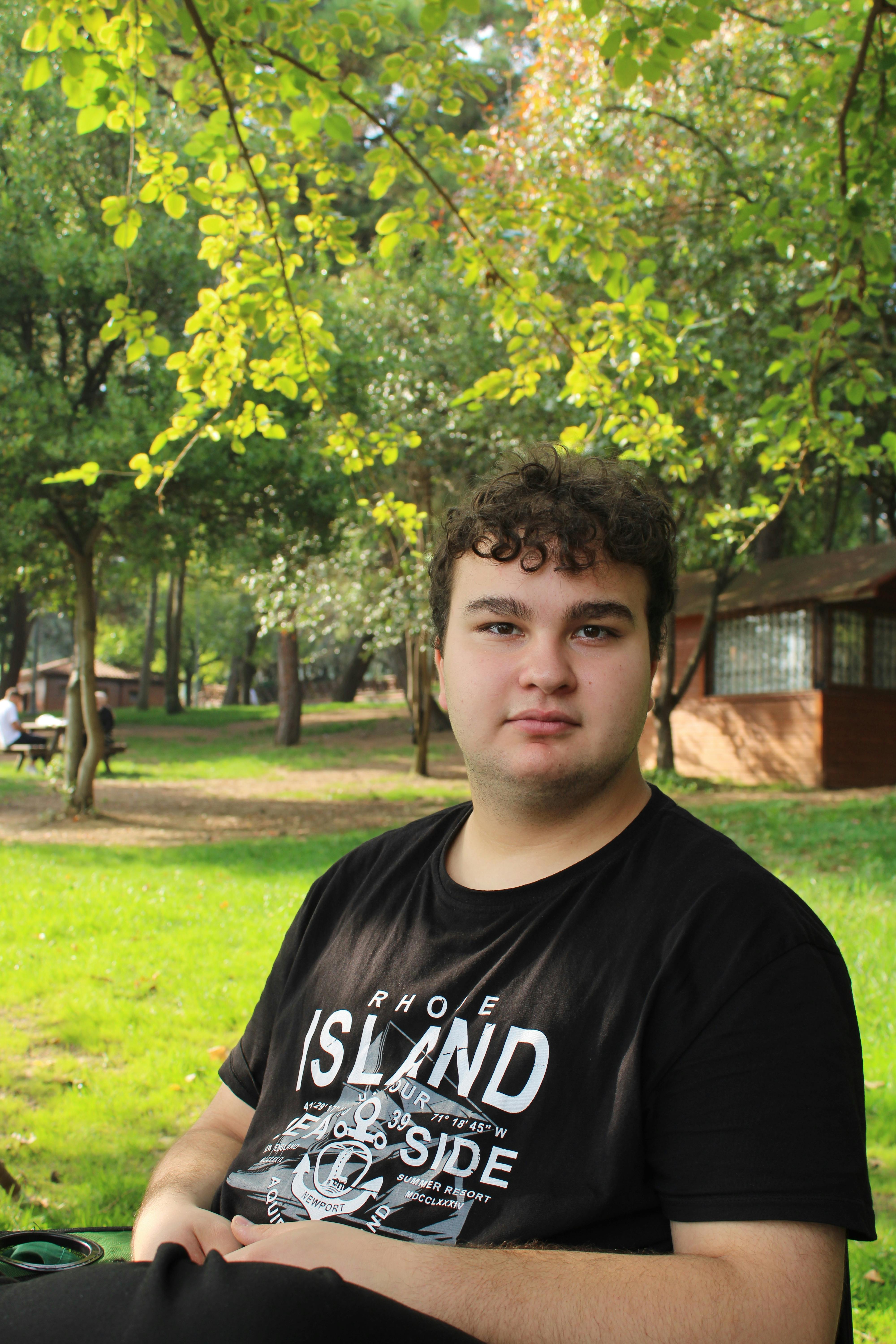 Young man sitting on a bench in a green park surrounded by trees and a cabin.