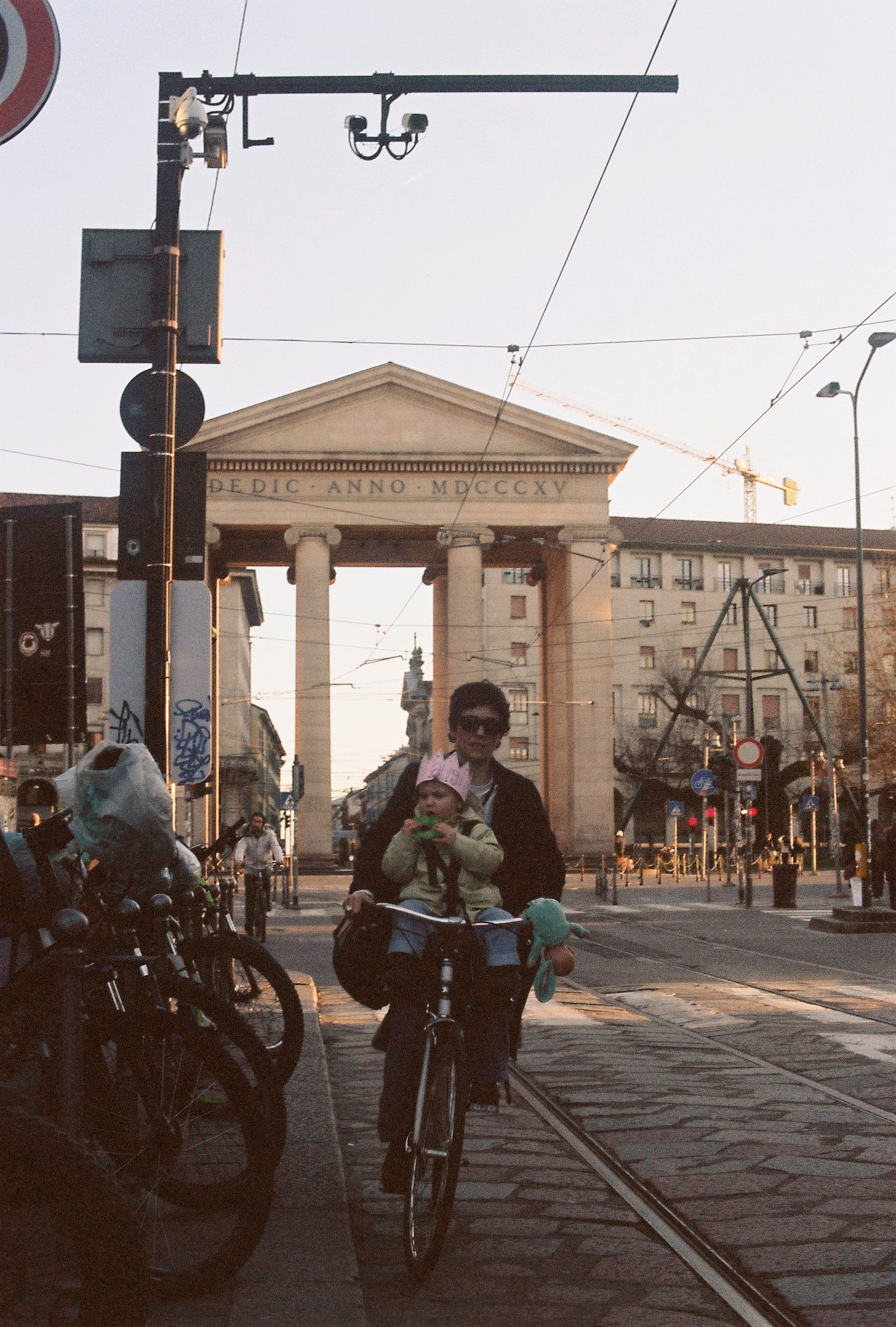 Cyclist with child rides past Milan's Porta Ticinese on a sunny day, capturing urban life.