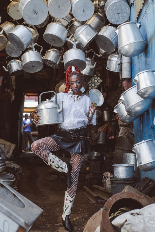 Free A fashionable individual poses creatively amongst hanging metal pots in Nairobi's vibrant market. Stock Photo