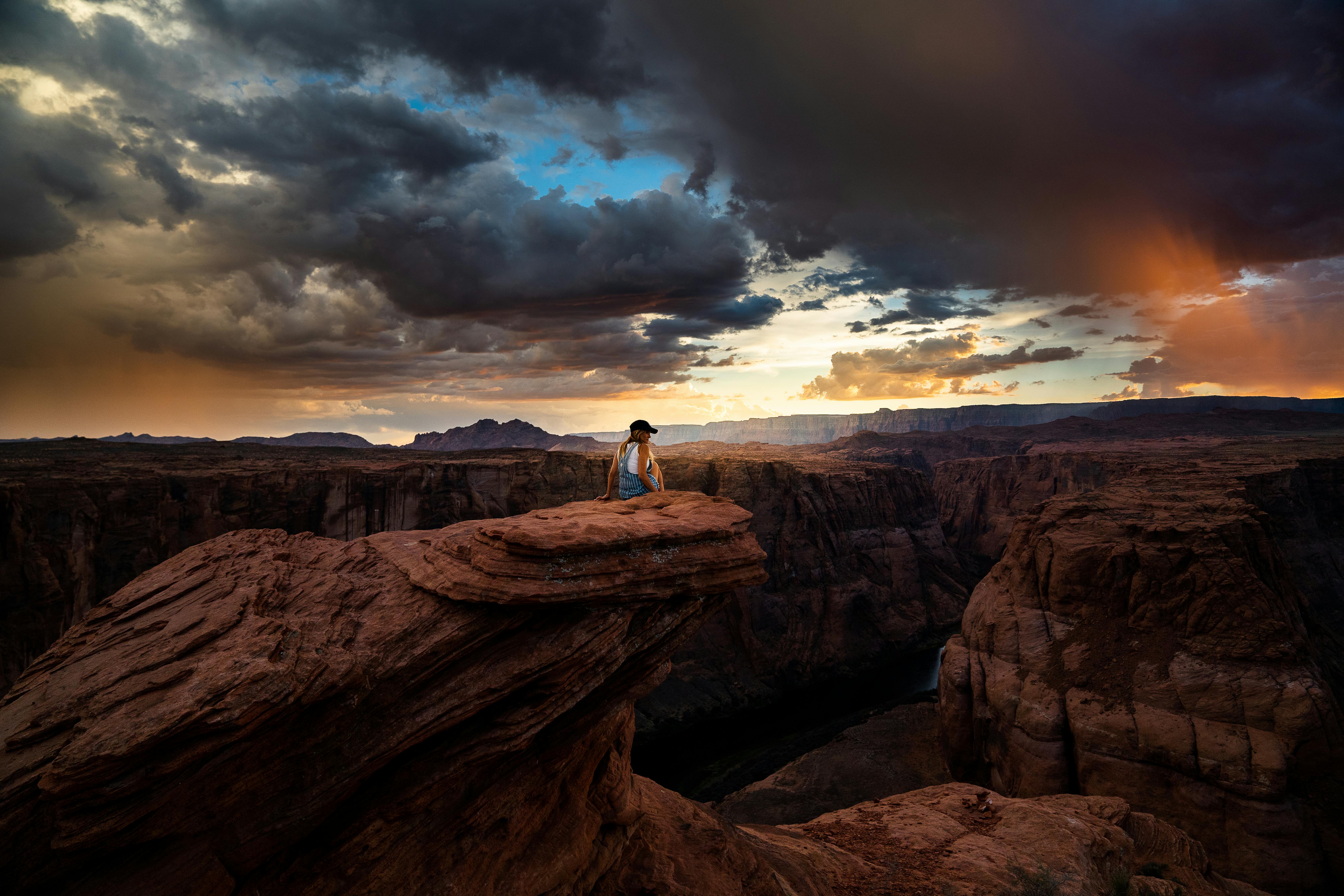 Person sitting on cliff at Horseshoe Bend during a dramatic sunset in Arizona.