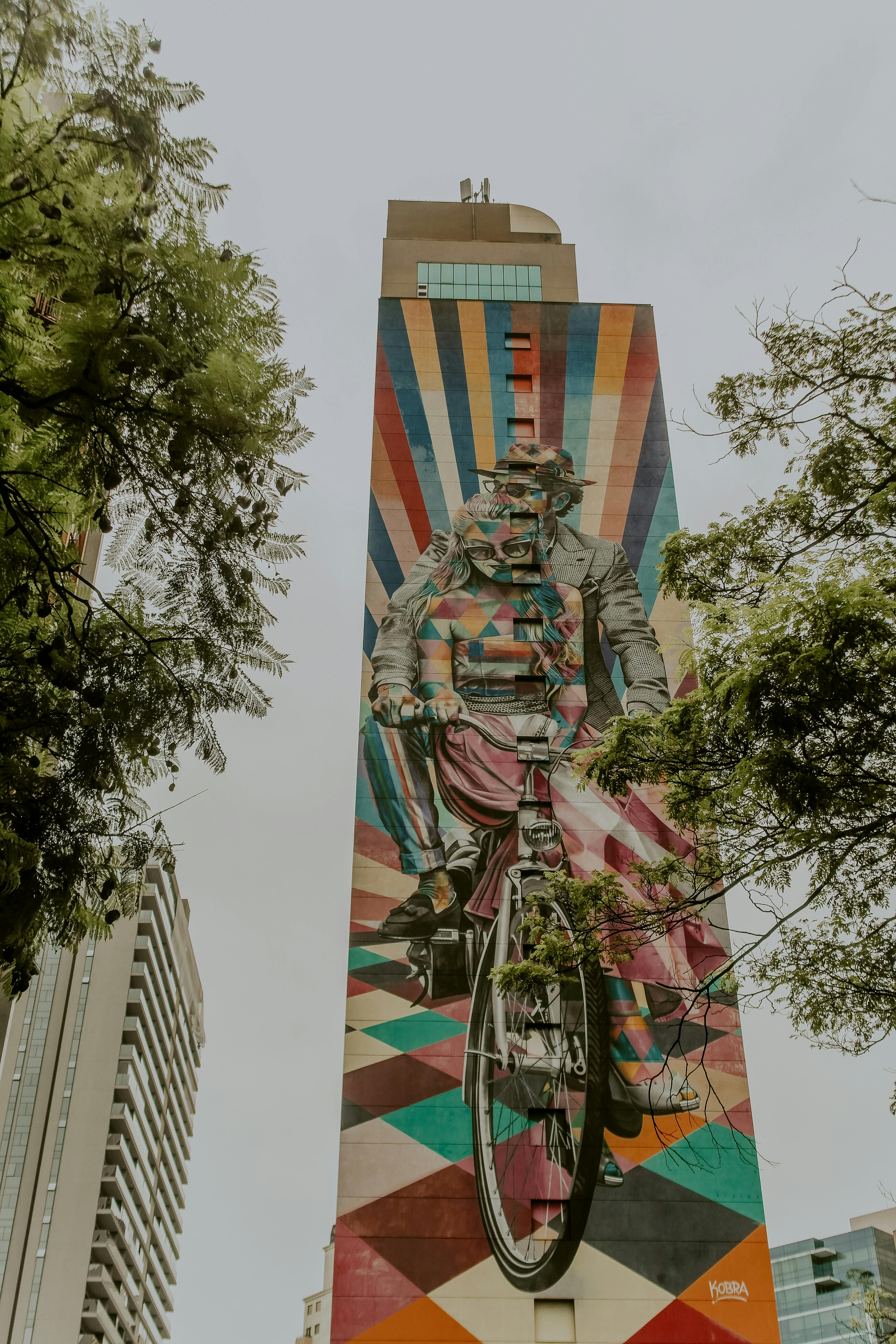 一幅色彩缤纷的壁画描绘了城市高楼之上，一位骑自行车的人穿行于建筑与绿荫之间的景象