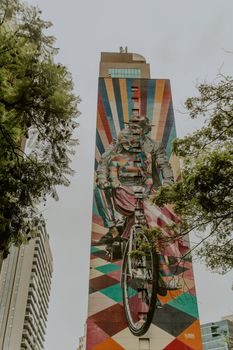 一幅色彩缤纷的壁画描绘了城市高楼之上，一位骑自行车的人穿行于建筑与绿荫之间的景象