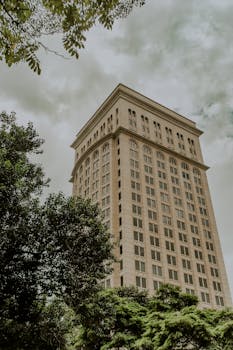 多云天空下，一座被绿色植物包围的高大摩天大楼映入眼帘