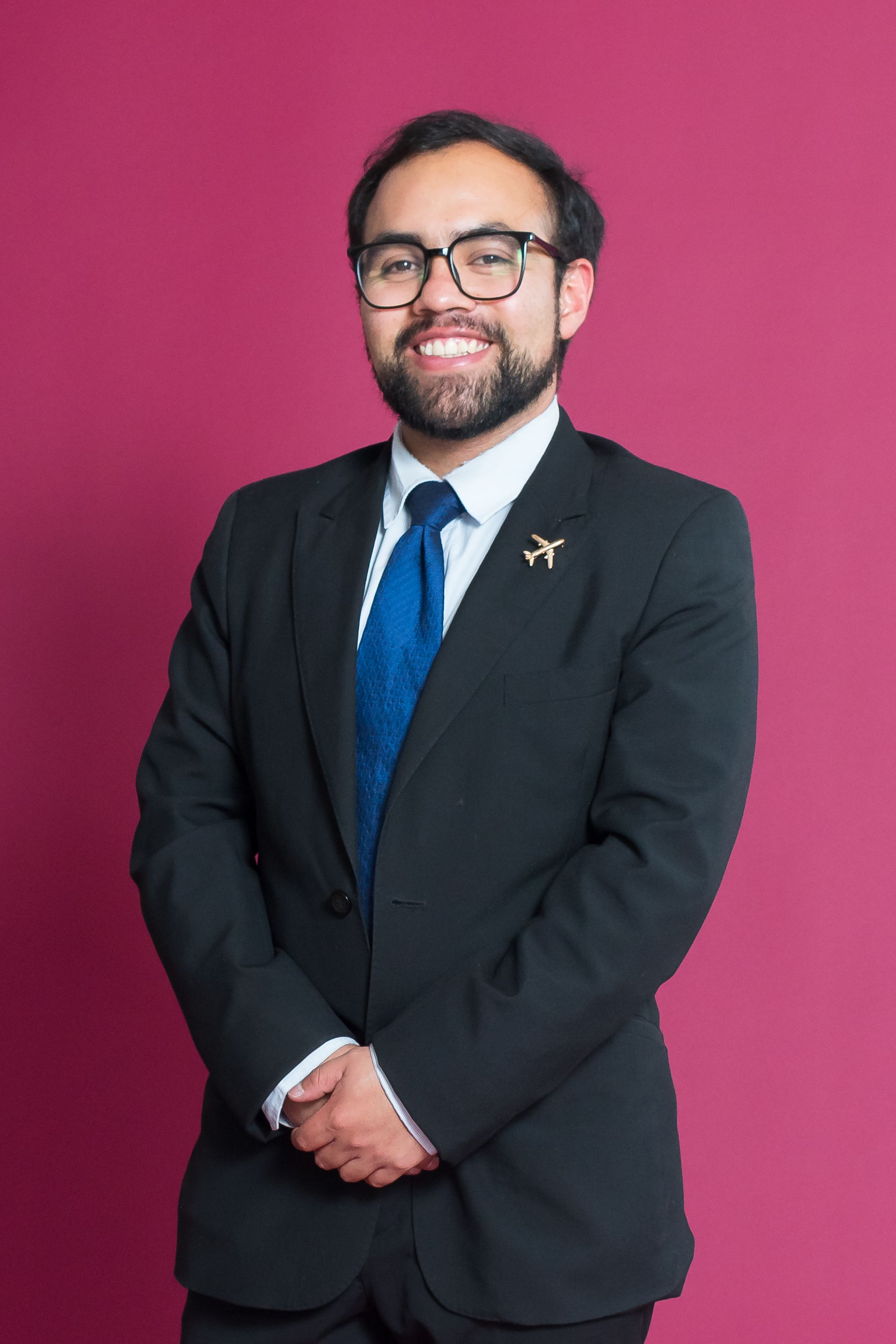 Confident businessman in a suit and airplane pin stands against a magenta background.