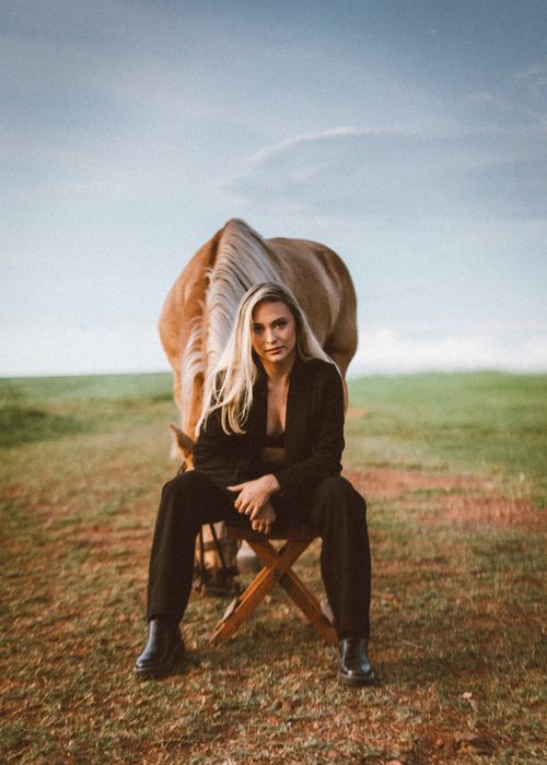 Free Blonde woman seated on a chair in the countryside with a brown horse behind her. Stock Photo