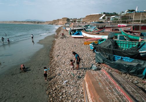 Children playing near colorful boats on a pebbled beach in Pacasmayo, Peru, under a bright day sky.