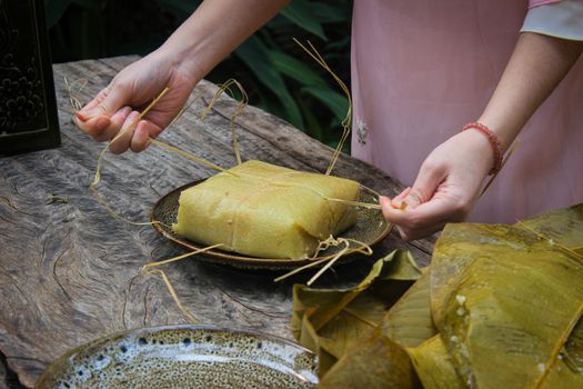 Close-up of hands wrapping a traditional Asian rice dish with leaves on a rustic table.