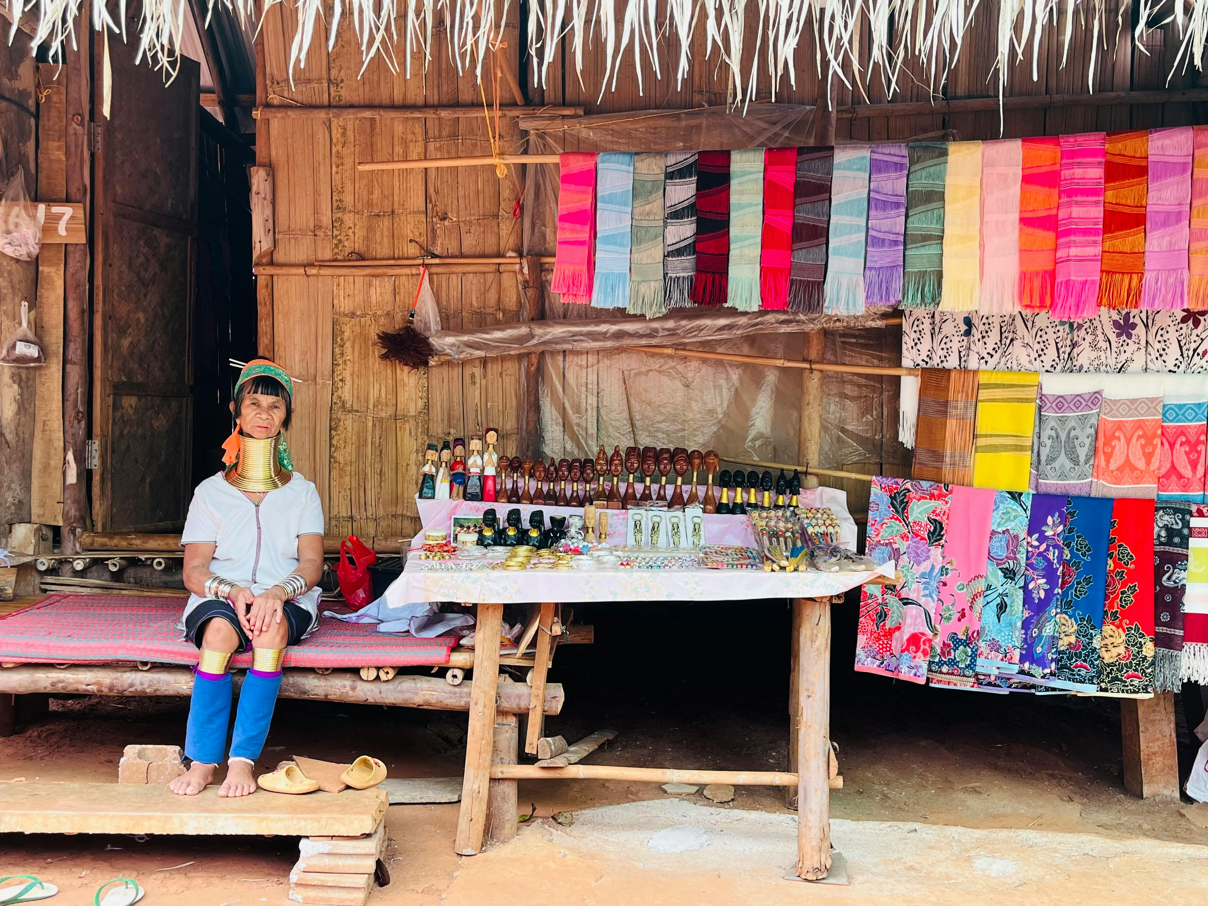 A Karen woman in traditional attire sits at a vibrant textile market in Chiang Rai, Thailand.