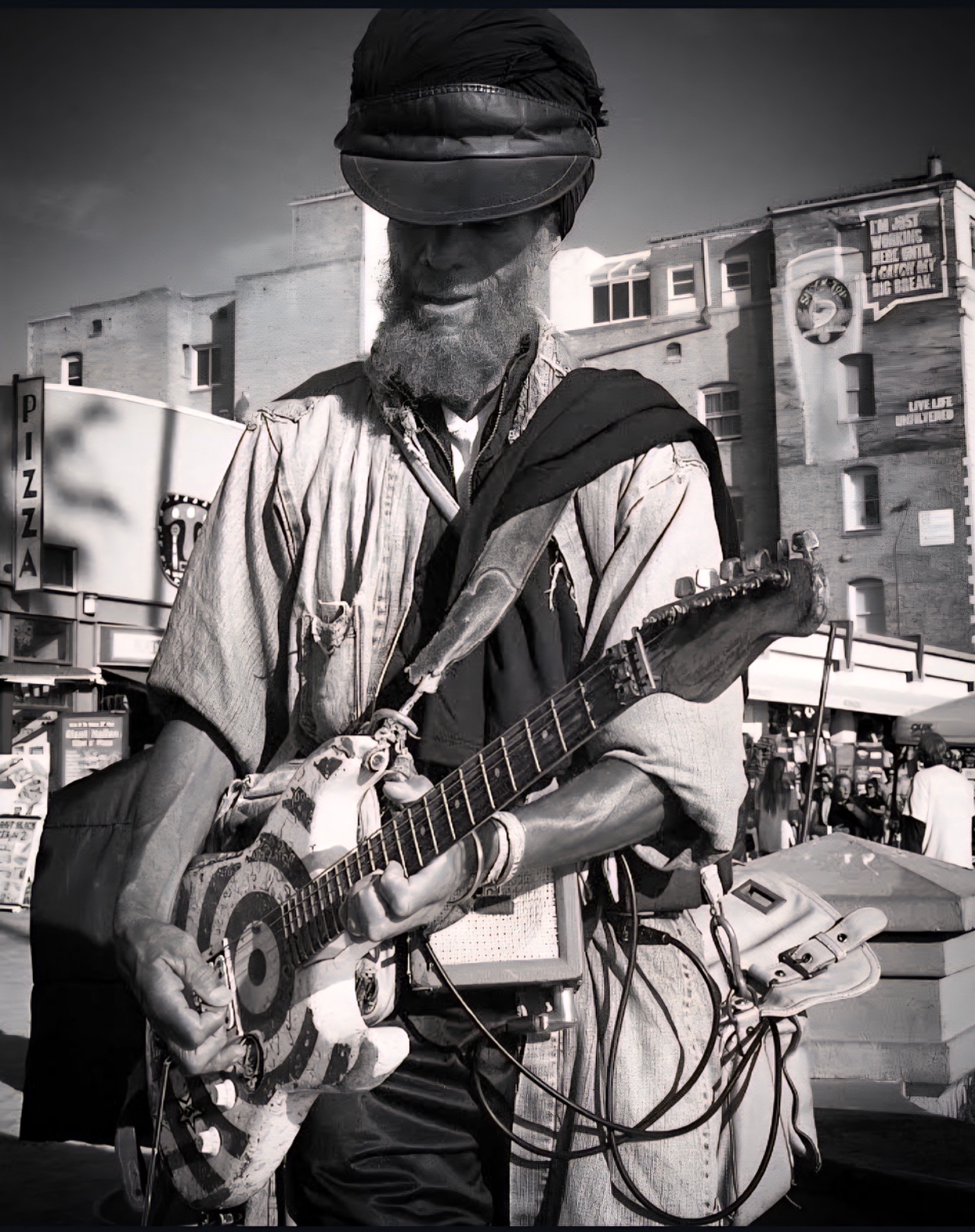 Black and white photo of a street musician playing guitar outdoors in an urban area.
