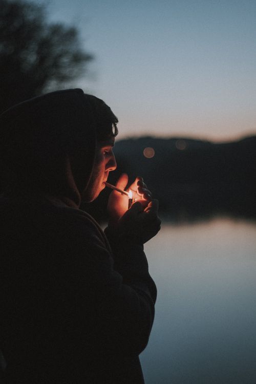 Free A man lights a cigarette by a calm lake during a serene evening sunset, creating a moody silhouette. Stock Photo