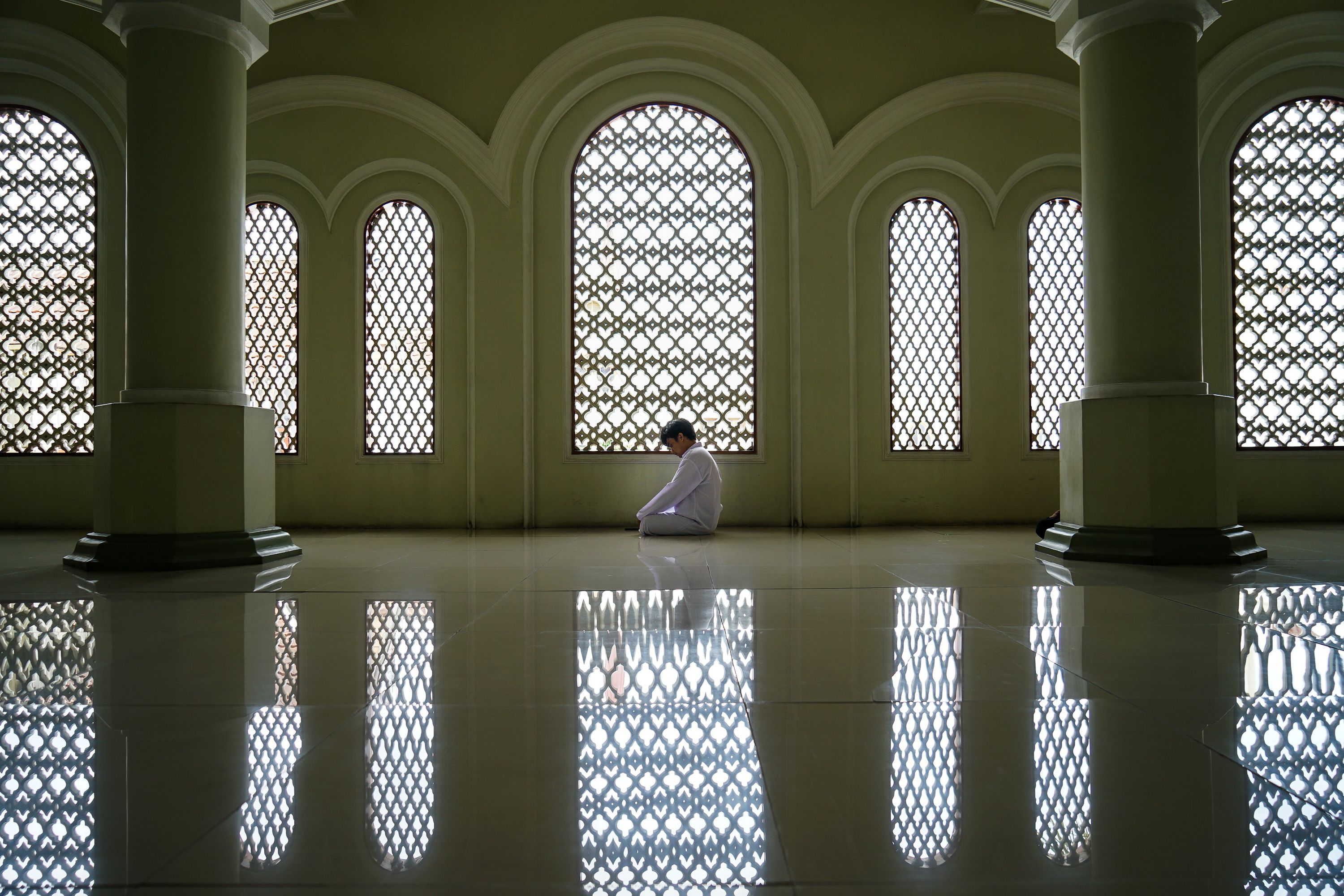 A person seated in meditation in a beautifully arched hallway with reflective flooring and intricate window designs.