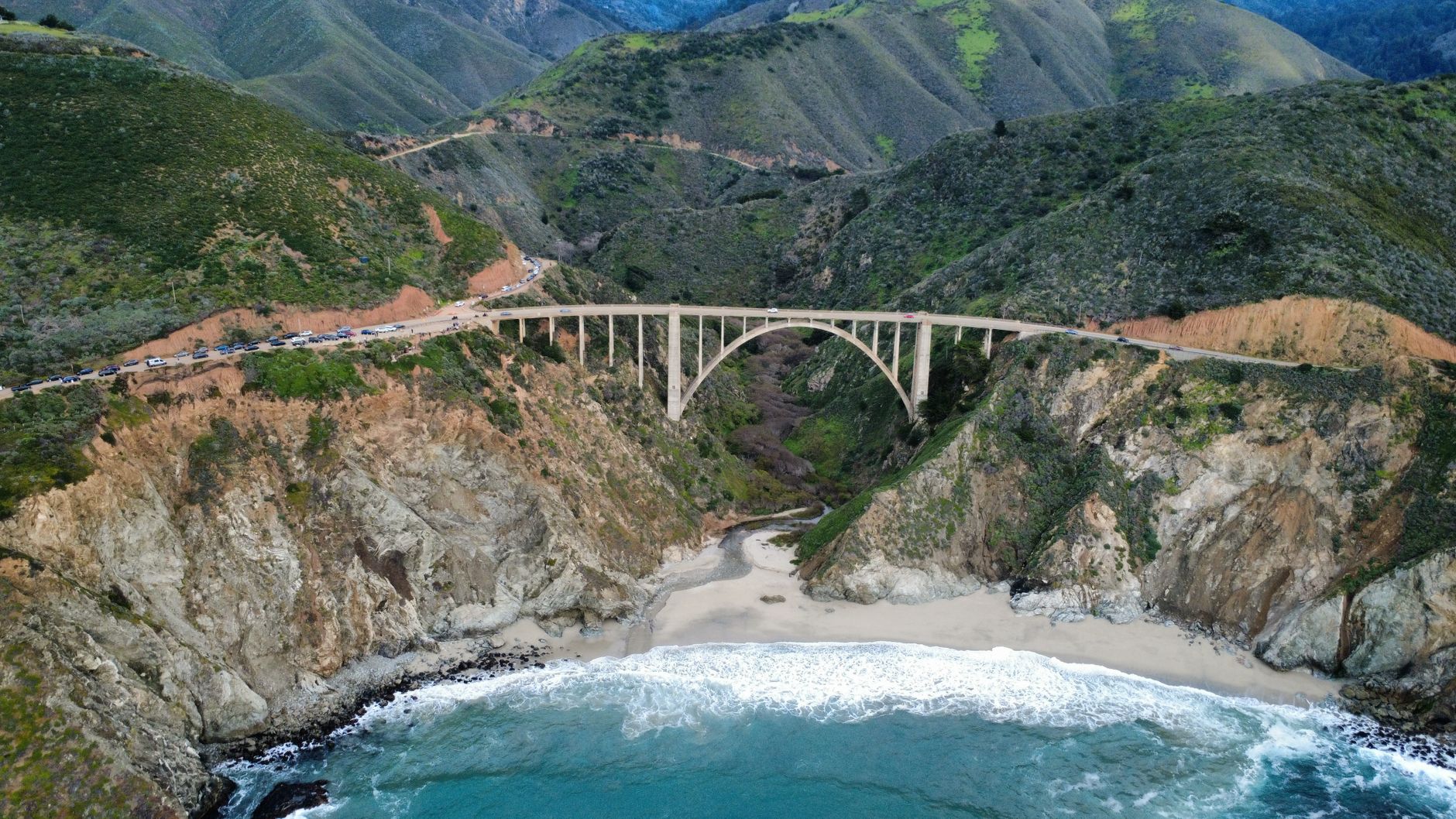 Aerial view of Bixby Creek Bridge along Big Sur coast with lush hills and the Pacific Ocean.