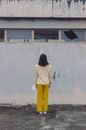 一名穿着黄色裤子的妇女站在风化的城市墙壁上，从背后被拍摄
