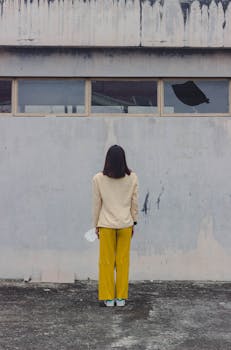 一名穿着黄色裤子的妇女站在风化的城市墙壁上，从背后被拍摄