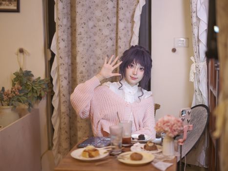 A woman in a cozy cafe, wearing a pink sweater, waves while sitting at a table with food and flowers.