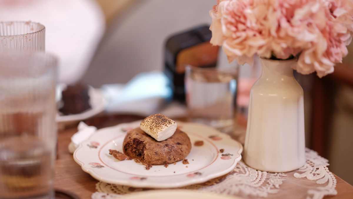 Elegant dessert scene with a chocolate cookie and marshmallow alongside pink flowers indoors.