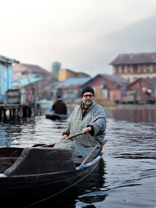 Free A man wearing winter clothes rows a wooden boat on a calm lake surrounded by colorful houses. Stock Photo