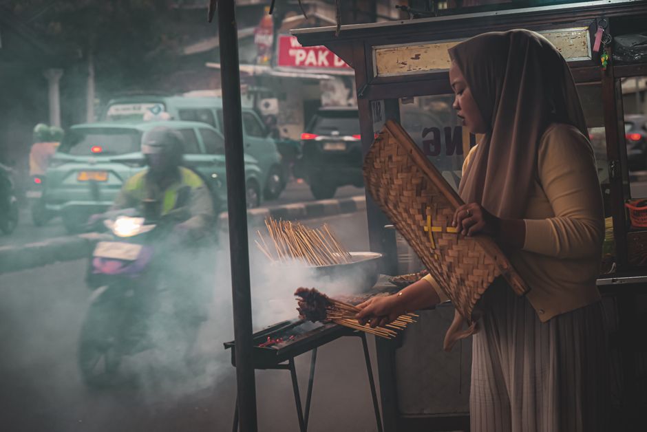 A woman grilling satay at a street stall in Jakarta, Indonesia, during the evening.