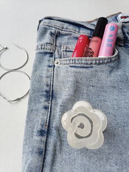 Blue denim jeans with lip products in a pocket and hoop earrings beside them.