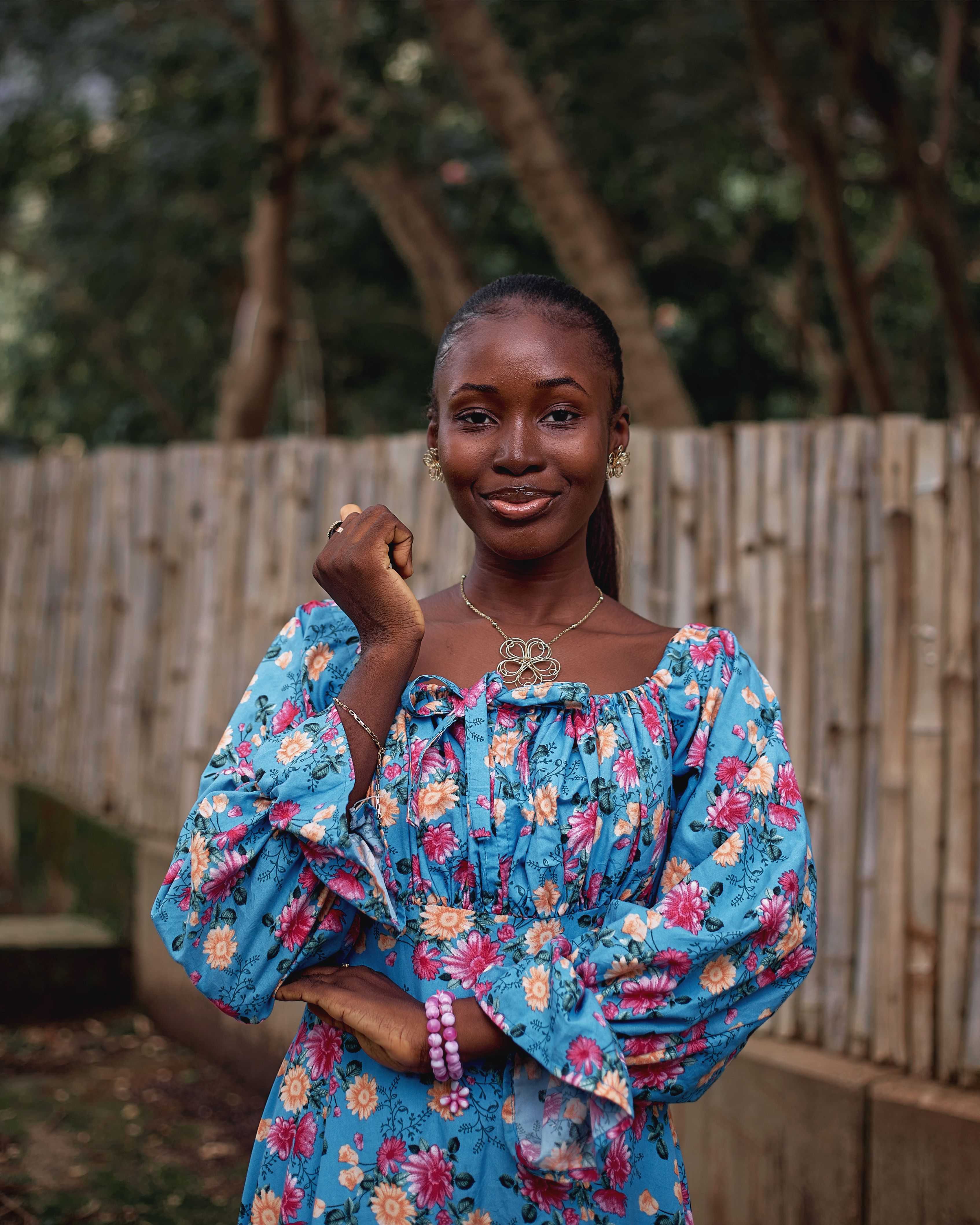 A young woman in a colorful floral dress stands smiling outdoors against a wooden fence backdrop.