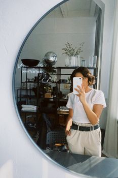 Young woman takes a selfie in a modern, minimalist studio setting with a large mirror reflection.